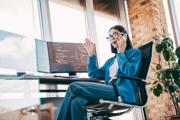 Businesswoman coder on dual monitors in a bright loft office talks on smartphone during coding and programming work
