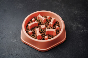Pink pet bowl with slow feeder maze filled with brown kibble on dark stone surface