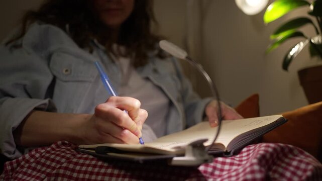 Close-up of a woman's hands capturing her innermost thoughts in a journal with a pen in the quietness of the night, nestled in a snugly lit room with a soft reading light casting a warm glow