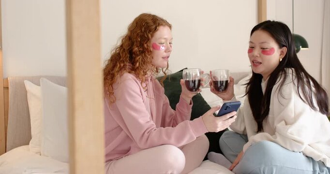 Diverse female friends sitting on bed wearing gel patches, reading smartphone and clinking glasses