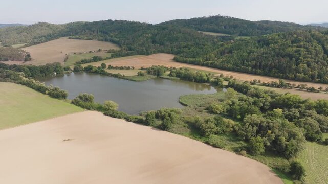 Aerial view of a serene lake reflecting the sky, bordered by lush green trees and harvested fields, creating a beautiful contrast, Chotilsko, Central Bohemian Region, Czechia.