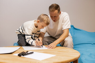 Obraz premium Father Teaching Son How to Draw at Home While Sitting at Wooden Table in Cozy Modern Interior, Concept of Creative Learning and Family Bonding