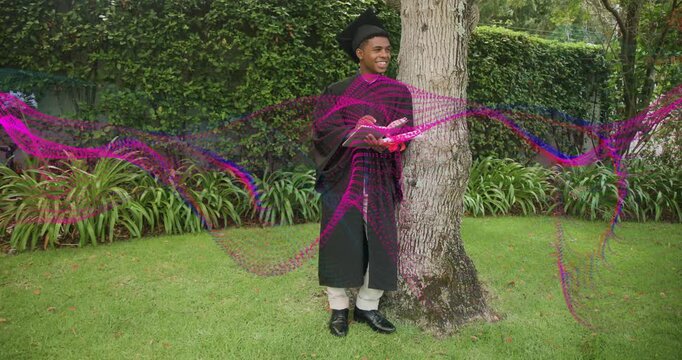 Graduate walking to tree, leaning, signing graduation booklet while pink ribbon sweeping over him