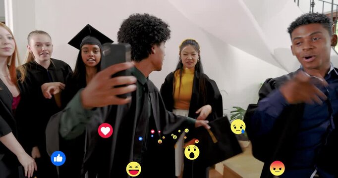 Man raising phone, graduates handing caps and hugging, emoji overlaying, posing graduation selfie