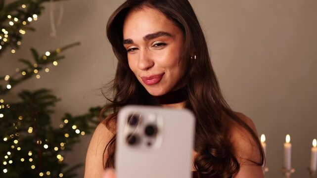 Close-up of an attractive woman in a festive velvet outfit smiling and adjusting her hair while participating in a video call or recording a vlog, with a glowing Christmas tree background.