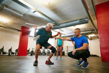 Fototapeta premium Coach, old man or squat in gym class with pipe for mobility, fitness activity or exercise instructions. Personal trainer, senior person or pvc equipment with help for workout, stability or low angle.