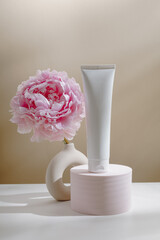 White mockup face cream tube on podium with pink peony flower in vase and aesthetic shadows, minimal neutral tone. Skincare mockup bottle, body lotion, feminine moisturizer product