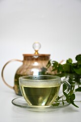 A cup of mint tea served on a glass saucer, surrounded by fresh leaves and a sophisticated teapot. A bright, calm photo, suitable for tea branding, health blogs, organic products