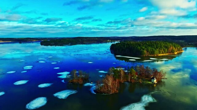 Aerial view of scattered ice floes on a tranquil lake, reflecting the cloudy sky, with islands covered in dense green forests, Mariefred, Sodermanland County, Sweden.