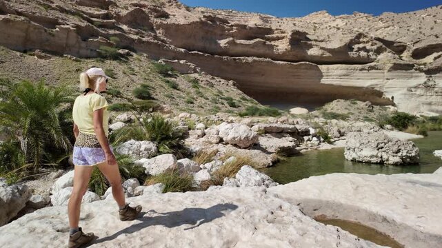 Woman actively trekking through the rocky terrain of Wadi Hadhbram canyon in Oman, navigating across large boulders with palm trees and a small stream in the natural desert oasis in Dhofar