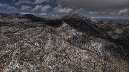 Aerial view of the winding, dark ribbon of a road cutting through the snow-dusted, rocky slopes of the mountains under a sky streaked with clouds, Tucson, Arizona, United States.