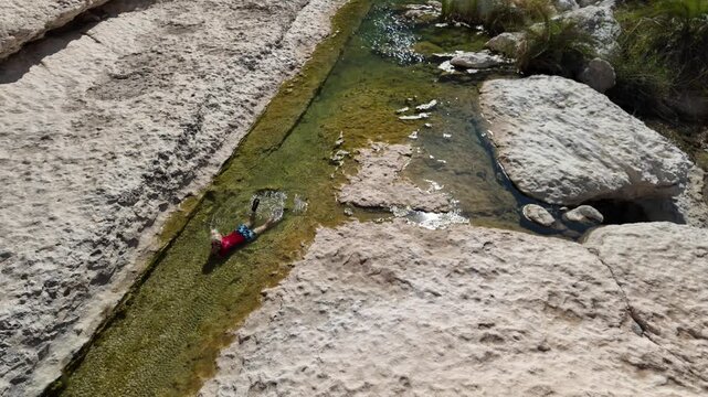aerial view of girl floating peacefully in a clear green natural pool in Wadi Hadhbram, Oman, enjoying a refreshing swim amid rocky canyon walls and sunlit tranquil desert waters in Dhofar