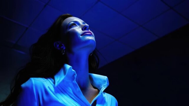 A woman in a white blouse gazes upward under blue lighting, with a dark ceiling grid in the background.