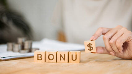 Hand putting wooden block with dollar sign to complete word BONUS on table.