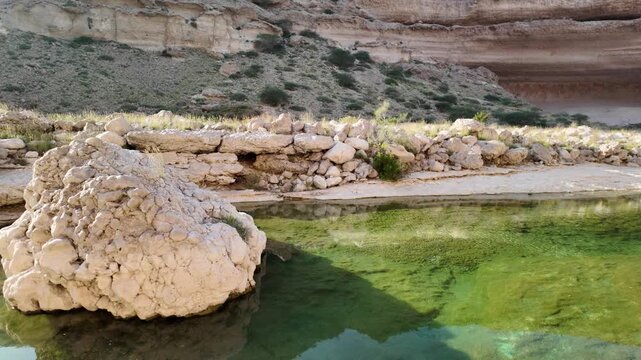 Wadi Hadhbram in Oman presenting a serene desert oasis with clear green water reflecting the surrounding rocky mountains, boulders, and sparse vegetation under the warm daylight in Dhofar