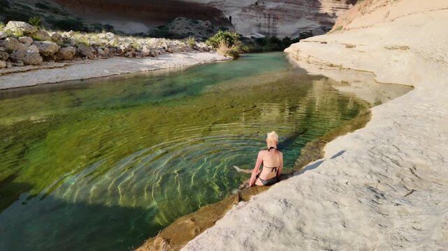 Woman sitting at the edge of a natural pool in Wadi Hadhbram in Oman, bathing in clear green water surrounded by rugged canyon walls, finding serenity and escape in the tranquil desert oasis in Dhofar