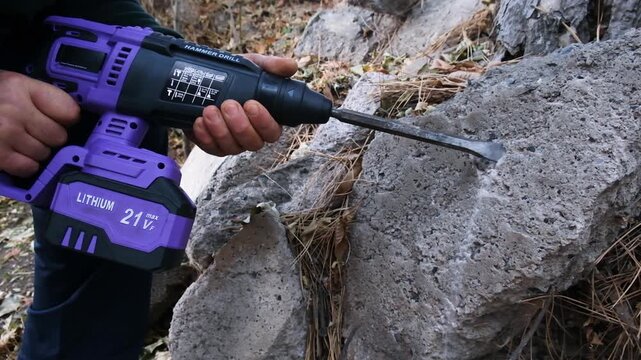 Low angle shot from the ground: a man is using a purple hammer drill to chisel concrete outdoors. A flat metal chisel bit for hard stone is inserted into the drill. Concrete demolition.