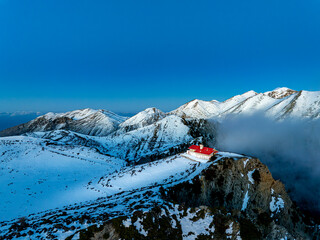 Aerial view of a solitary red-roofed building atop a snow-dusted peak amidst the rugged, cloud-kissed mountains under a vibrant blue sky, Omalos, Chania, Greece.