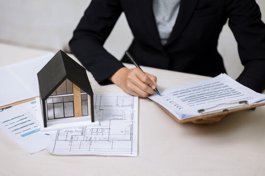 Real estate agent writing on document checklist with house model and blueprints.