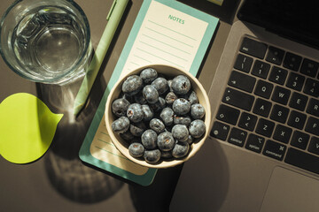 Bowl of fresh blueberries and glass of water on office desk with laptop concept of workplace...