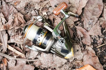 A metal fishing reel with a wooden handle placed on the ground with dried leaves by the riverbank.