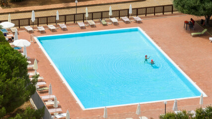 Aerial view of a swimming pool with two people swimming. The pool is in the garden of a hotel. There are many sun loungers and deckchairs by the pool. 