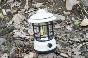 A white portable hanging electric camping lantern placed on soil and rocky ground during outdoor activities.