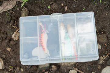 A small transparent fishing tackle box containing lures, hooks, bobber, and sinker, placed on the ground by the riverbank.