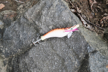 An orange shrimp-shaped fishing lure used for squid and cuttlefish placed on a rocky surface during fishing activities.