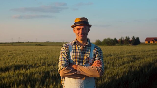 Portrait of smiling satisfied farmer whit hat standing in wheat field at sunset looking at camera.