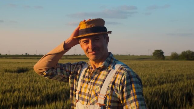 Portrait of smiling satisfied farmer whit hat standing in wheat field at sunset looking at camera.