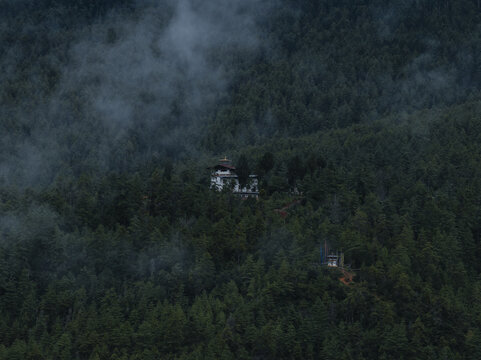 Aerial view of a monastery nestled amidst the dense, verdant forest, with mist swirling around the treetops, Paro, Bhutan.
