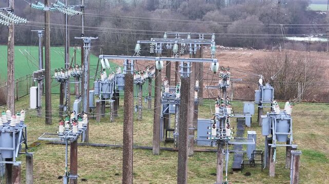 A fenced rural electrical substation surrounded by power lines, transformers, open agricultural fields and distant forest under an overcast sky, expressing industrial presence in natural countryside.