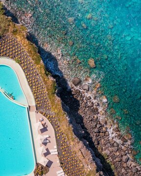 Aerial view of rocky cliffs meeting the turquoise sea next to a resort with pools and rows of beach chairs, Vulcano, Sicily, Italy.