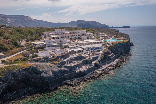 Aerial view of a white hotel complex perched on a rugged cliffside overlooking the turquoise sea, with the volcanic landscape in the background, Vulcano, Sicily, Italy.