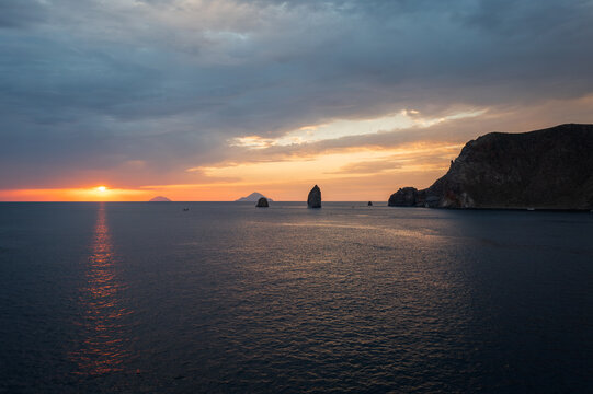 Aerial view of the sun sinking below the horizon, casting a warm golden glow over the tranquil waters near rocky outcrops, Vulcano, Sicily, Italy. - Powered by Adobe
