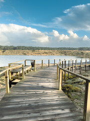Naklejka premium Wooden Boardwalk Path by Coastal Lagoon Ovar A wooden boardwalk path curves around a coastal lagoon in Furadouro Portugal leading towards a dense pine forest under a dramatic cloudy blue sky
