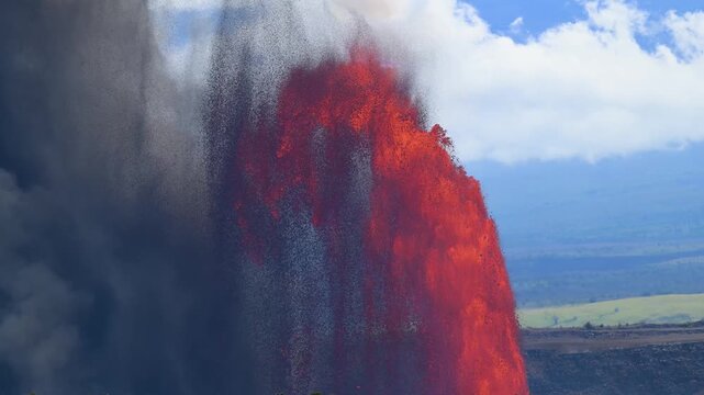 Aerial view of a dynamic volcano eruption, with vibrant red lava contrasting against the dark smoke and the green landscape, Kilauea, Hawaii, United States.