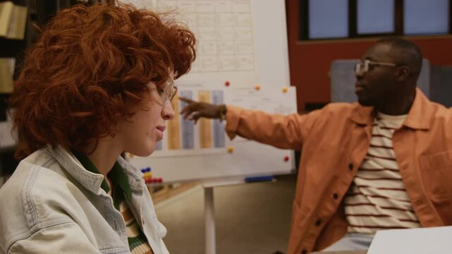Side view of curly red haired young woman writing grammar rules notes in English class while Black male teacher explaining new topic to group of students pointing at whiteboard in library