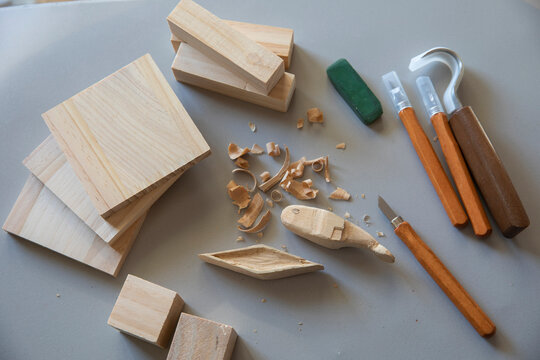 A top-down view of wood carving tools, including various chisels and knives, scattered among small wooden blanks and shavings, highlighting the creative hobby of woodworking.