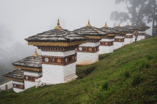 View of aligned Dochula Chorten's with white walls, grey roofs, and golden accents stand on a vibrant green hillside under a misty sky, Hungtso, Bhutan.