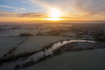 Aerial view of dawn's golden light kissing the River Thames, casting a soft glow over the misty fields and sleepy Sonning, Oxfordshire, United Kingdom.