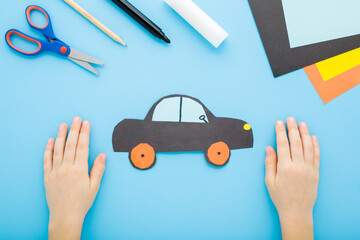Little child boy hands showing dark black car shape from application paper on light blue table background. Pastel color. Paper craft. Closeup. Point of view shot. Top down view.