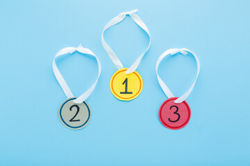 Created gold, silver and bronze paper medals with white ribbons on light blue table background. Pastel color. Closeup. Little child handcraft. Top down view.