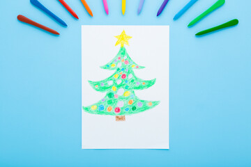 Wax crayons and drawn green Christmas tree with colorful baubles and yellow star on white paper on light blue table background. Pastel color. Closeup. Top down view.