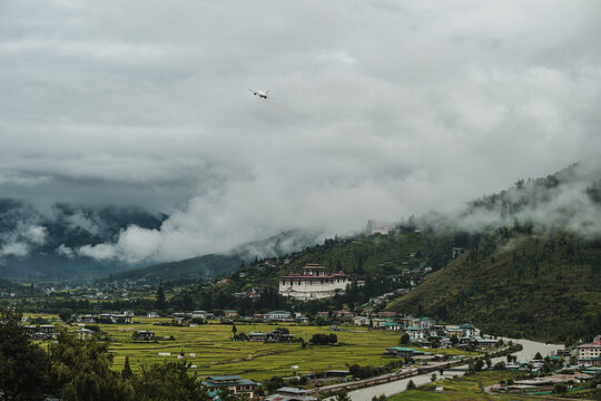 View of Rinpung Dzong nestled in misty mountains, where an airplane flies above the lush green valley, creating a serene scene, Paro, Bhutan.