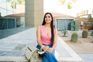 Young female latin student smiling looking away while sitting outdoors on university campus