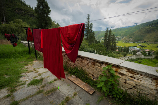 View of vibrant red robes hanging to dry against a backdrop of lush greenery and distant village under a cloudy sky, Paro, Bhutan.