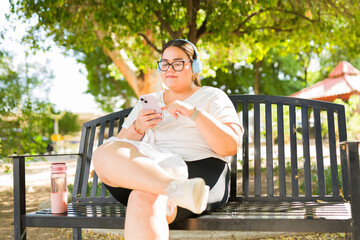 Fototapeta premium Overweight woman with headphones relaxing on a park bench, browsing smartphone and enjoying music outdoors on a sunny day