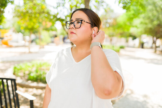 Woman listening to music or podcast with earbud and smartwatch in a park, enjoying leisure time outdoors - Powered by Adobe